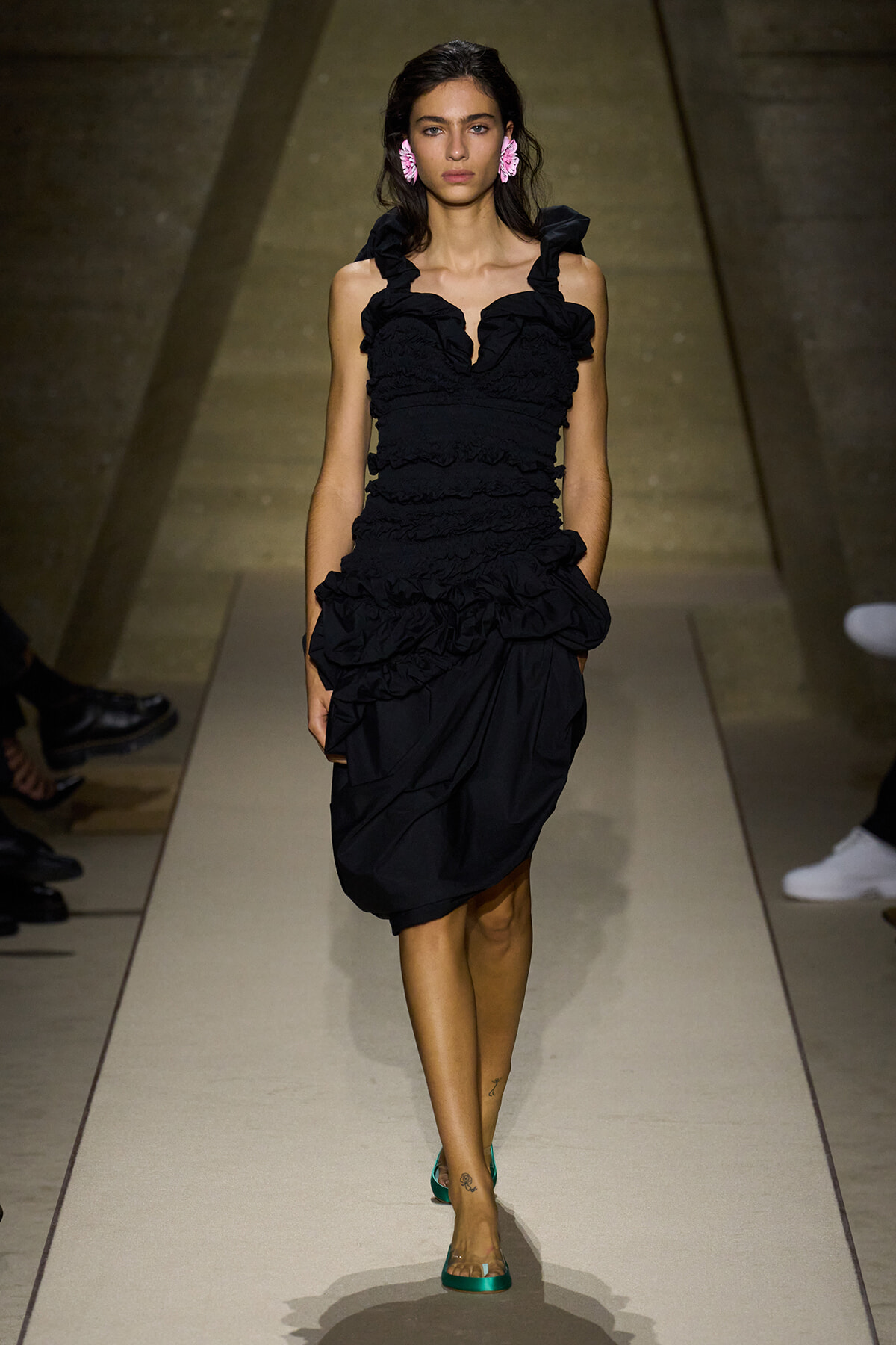 Female model walks a runway in a black ruffled dress with teal heels and pink flower earrings, spotlight on her.