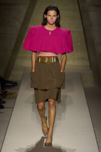 Model walking a runway in a bold fuchsia cropped blouse with puff sleeves and a wide tan layered skirt, cinched with a wide gold belt.
