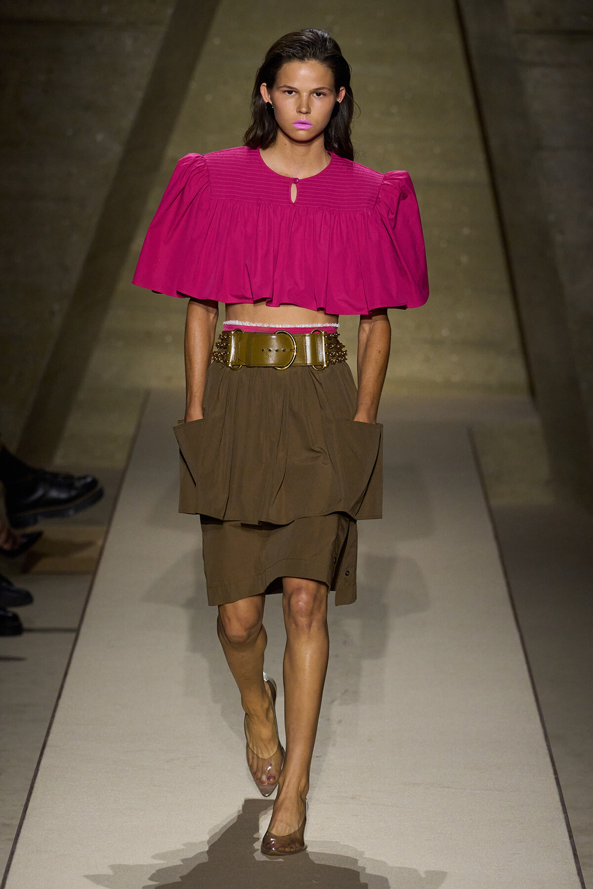 Model walking a runway in a bold fuchsia cropped blouse with puff sleeves and a wide tan layered skirt, cinched with a wide gold belt.