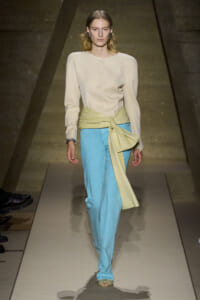 Model walking a runway in a cream long-sleeve, buttoned top with a pale yellow sash tied at the waist over light blue wide-leg pants.
