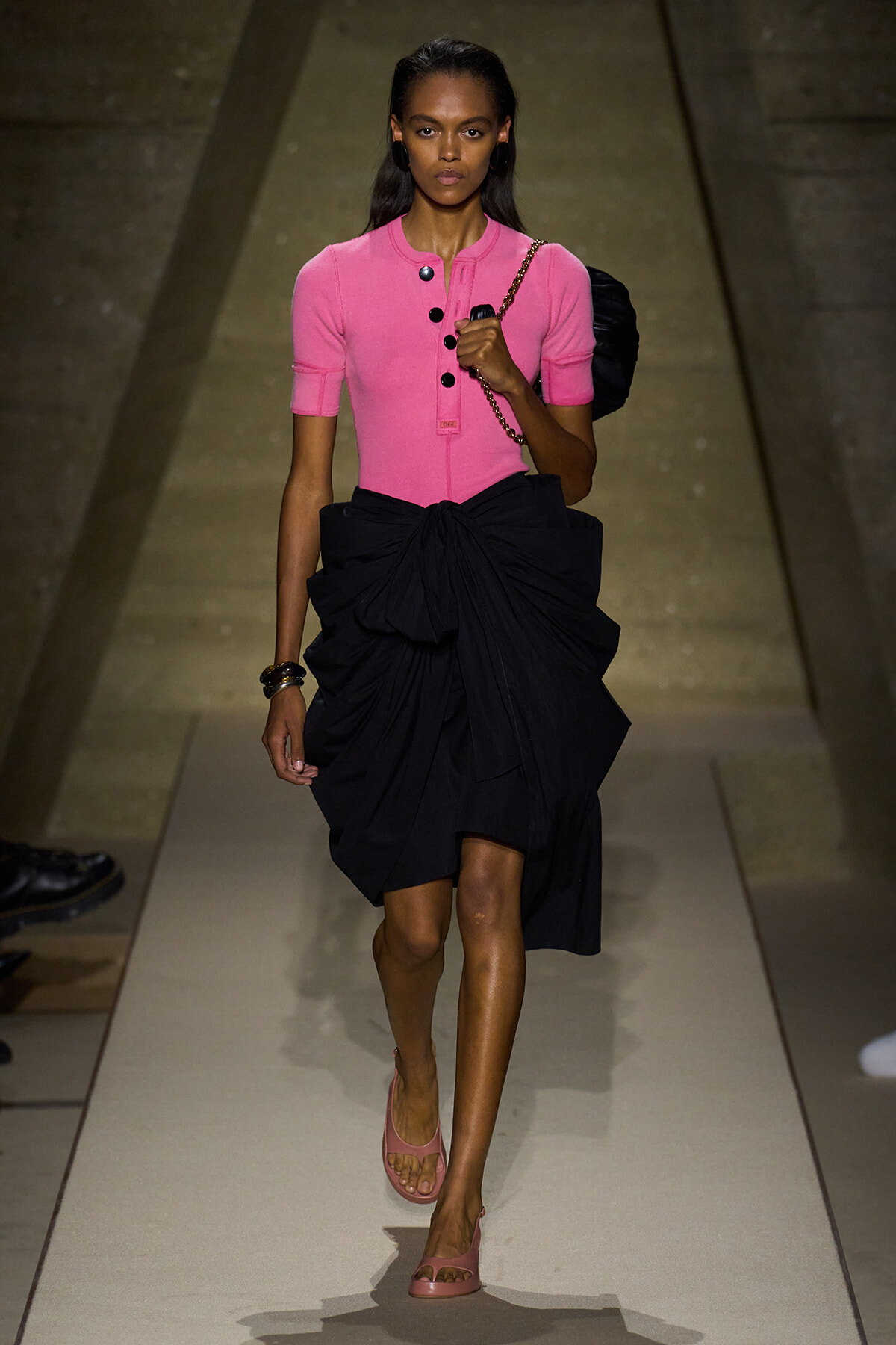Model walking a runway in a pink fitted top with a dramatic black bow skirt and a chain-strap bag slung over her shoulder.