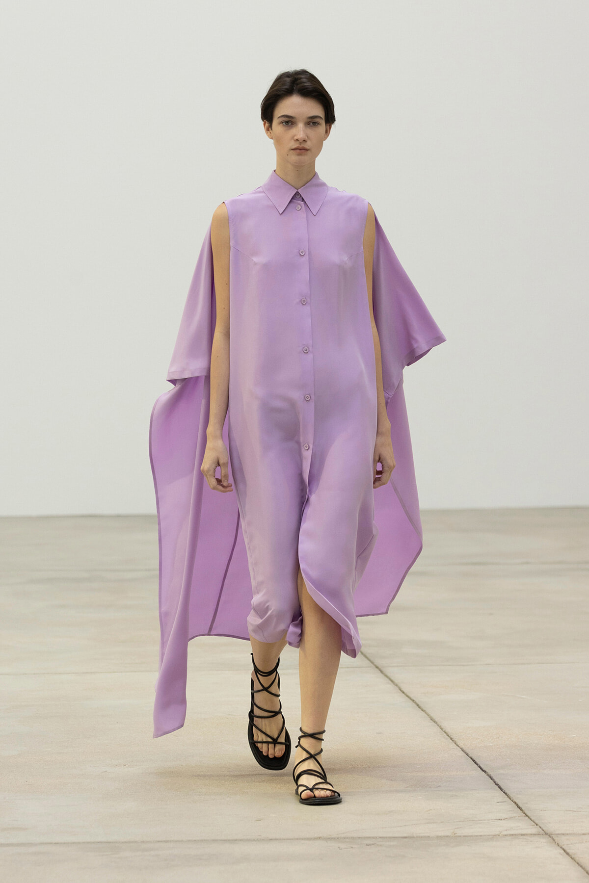 Model walking a runway in a lavender button-down dress with cape-like arms and black strappy sandals.
