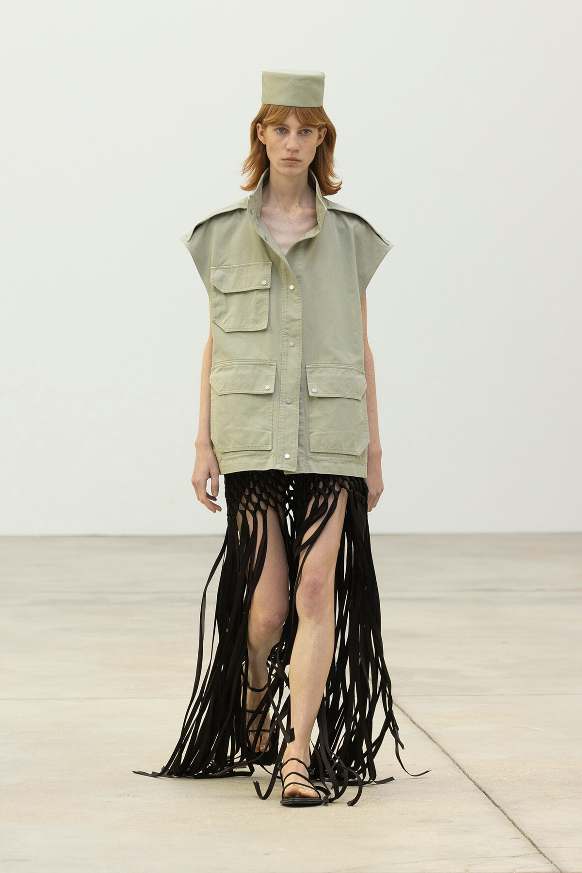 Model on a runway wearing a light green utility vest with multiple pockets, a matching square hat, and a long black fringe skirt with strappy sandals.