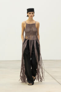 Runway model wearing a taupe lattice-knit top with long fringe strands over black wide-leg pants and a small black cap, walking forward.