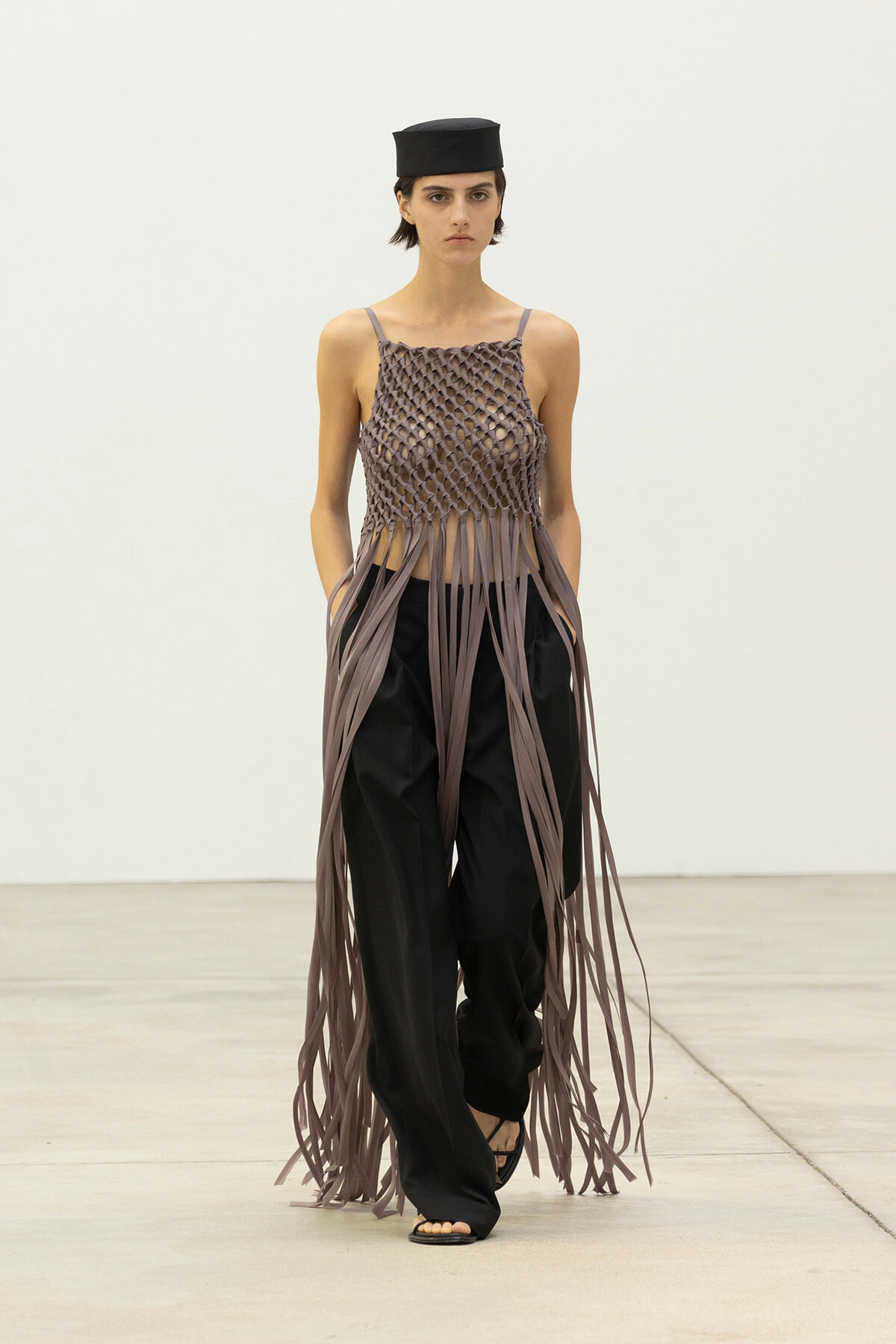 Runway model wearing a taupe lattice-knit top with long fringe strands over black wide-leg pants and a small black cap, walking forward.