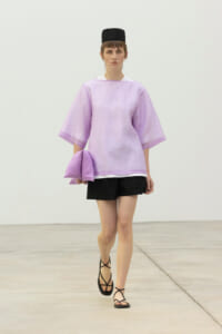 Model wearing a light purple oversized top with wide sleeves, black shorts, black sandals, and a black cap, carrying a matching lavender clutch on a minimalist runway.