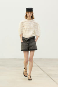 Model walking in a neutral studio wearing a white layered top, black denim shorts, strappy sandals, and a small black hat.