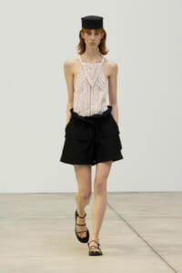 Model walking a runway in a pale pink crocheted halter top, black high-waisted shorts with large pockets, a small black hat, and strappy sandals.