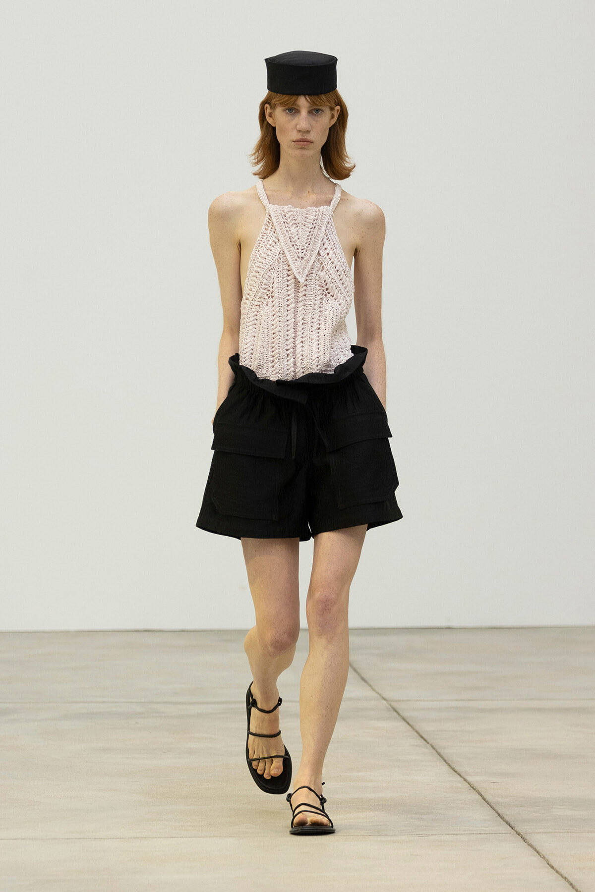 Model walking a runway in a pale pink crocheted halter top, black high-waisted shorts with large pockets, a small black hat, and strappy sandals.