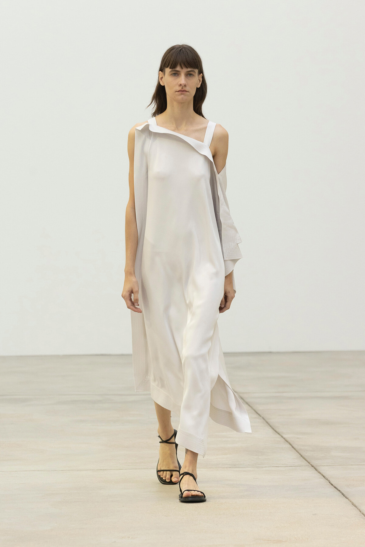 Female model walking on a runway in a white satin off-shoulder dress with a draped sleeve and black strappy sandals