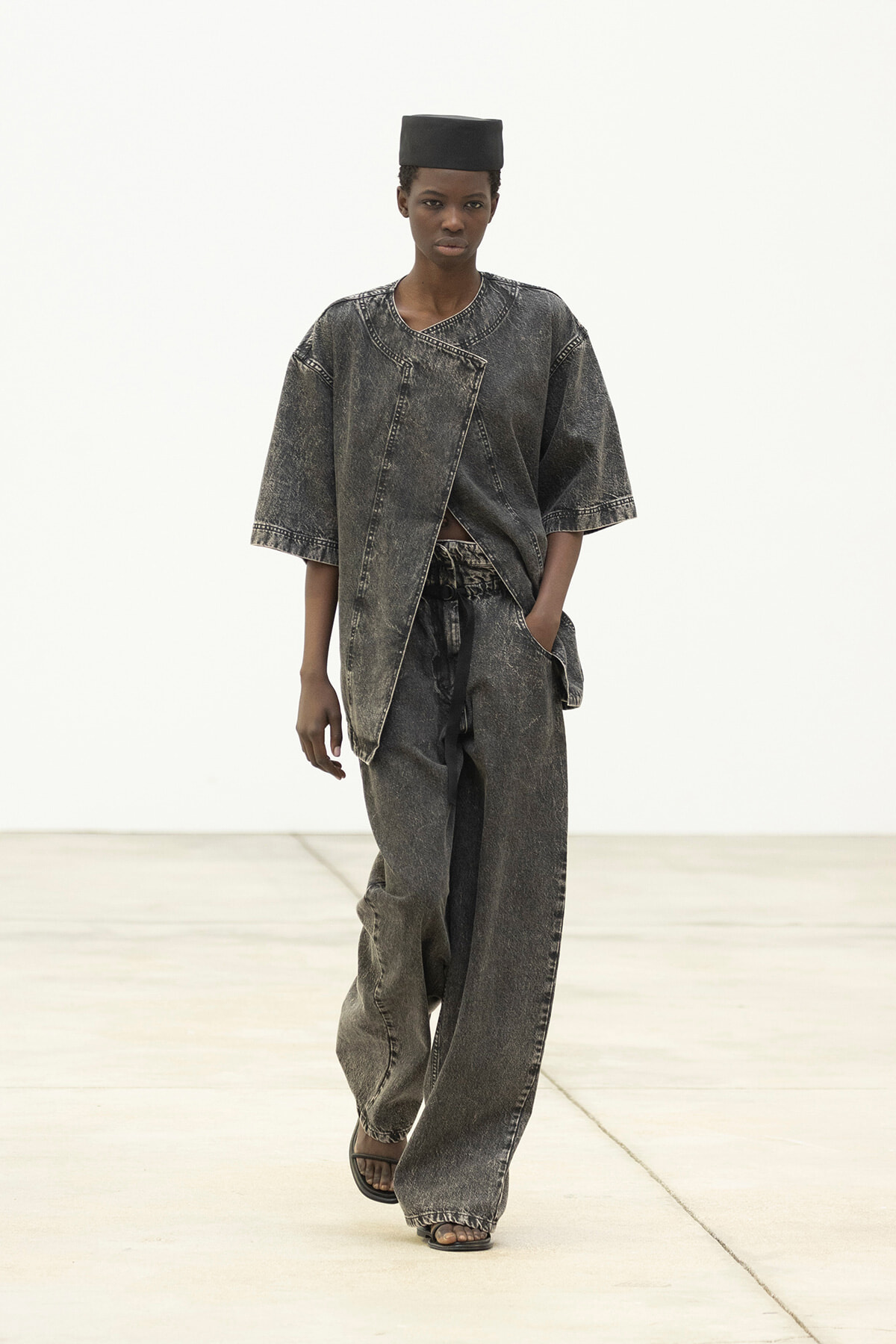Model walks in an oversized, acid-washed denim outfit: wide-leg pants and an asymmetrical top, with a black cap and sandals.