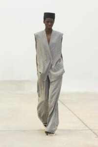 Model walking a fashion runway in a spacious, light setting, wearing a loose gray sleeveless double-breasted suit with wide-leg pants and a black square cap.