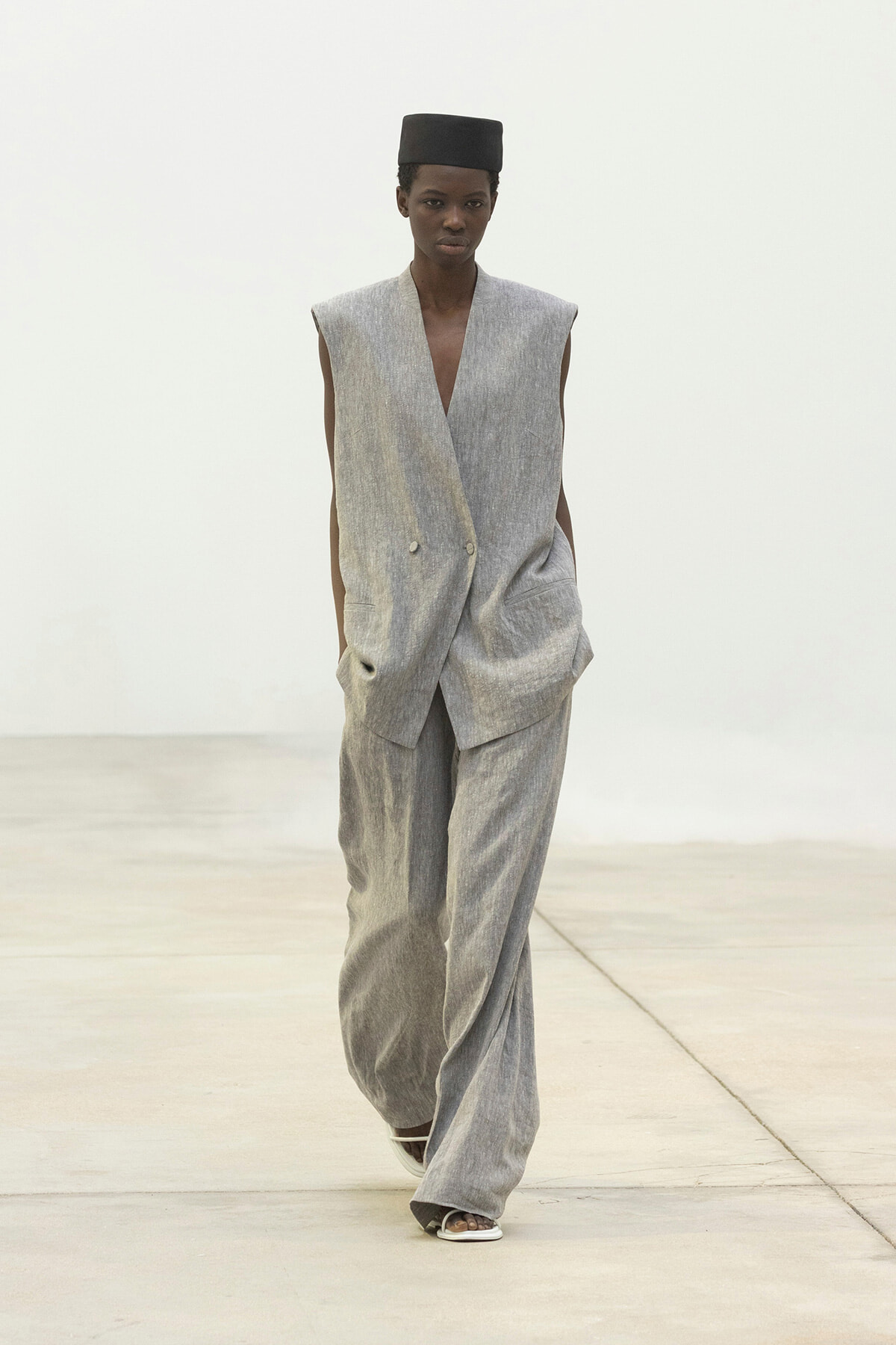 Model walking a fashion runway in a spacious, light setting, wearing a loose gray sleeveless double-breasted suit with wide-leg pants and a black square cap.