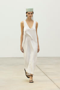 Model walking a runway in a white satin V-neck sleeveless dress with a gathered waist and black strappy sandals, plus a pale mint-green hat and long dangling earrings.