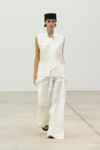 Model walking a runway in an all-white, sculptural outfit with a black cap.