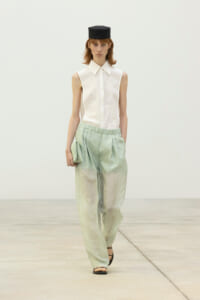 Runway model wearing a white sleeveless button-down top with pale green wide-leg pants and a small black hat, holding a matching clutch.