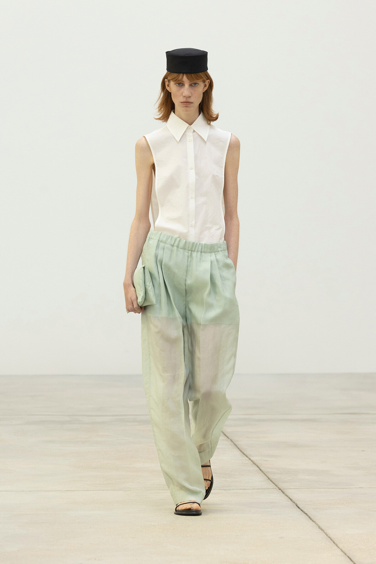Runway model wearing a white sleeveless button-down top with pale green wide-leg pants and a small black hat, holding a matching clutch.