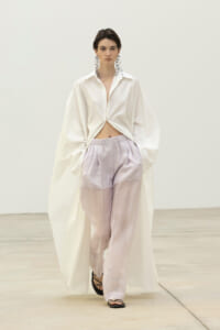 Model in a white knotted shirt over lavender sheer wide-leg pants, wearing dangling earrings and black sandals.