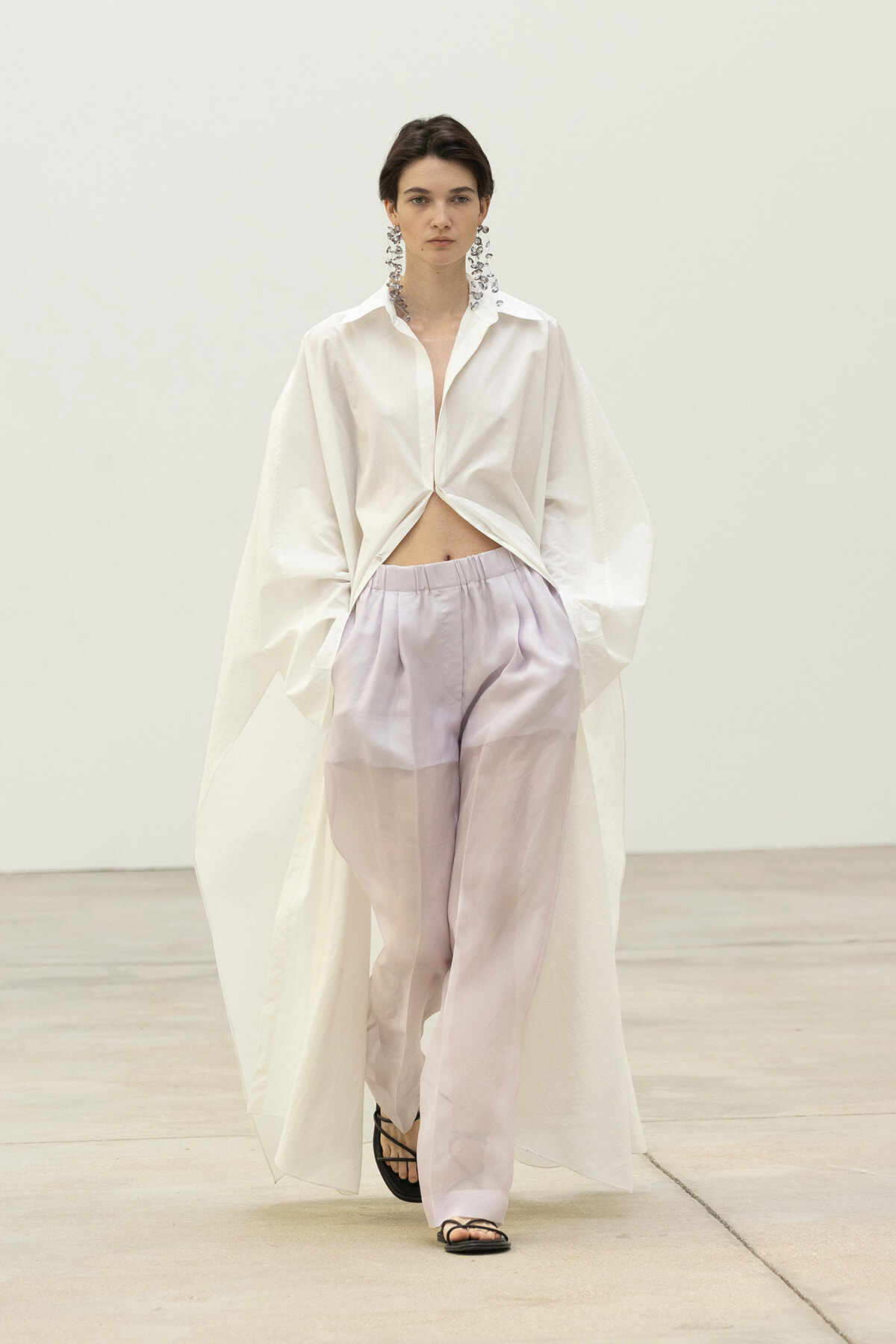 Model in a white knotted shirt over lavender sheer wide-leg pants, wearing dangling earrings and black sandals.