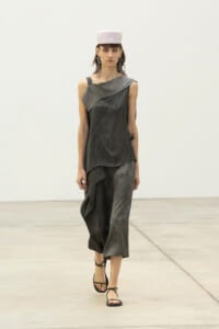 Runway model in a layered, dark gray draped dress with an asymmetrical hem, white hat, and strappy sandals.