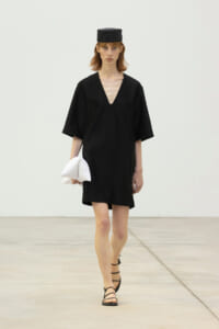 Runway model in a black V-neck dress with wide sleeves, black hat, strappy sandals, carrying a white folded clutch-like accessory.