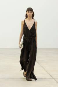 Runway model wearing a black V-neck sleeveless jumpsuit with a belt, holding a small patterned clutch