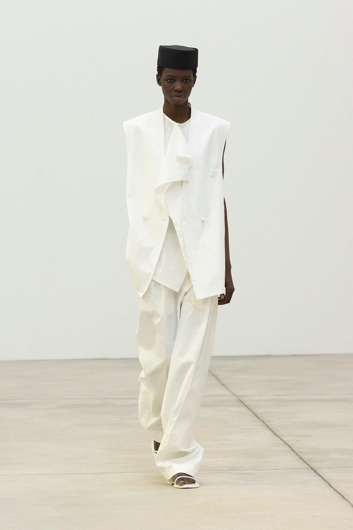 Runway look: an all-white, oversized suit with a draped front and wide-leg pants, paired with a black fez-style hat.