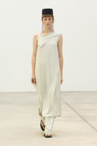 Model walking the runway in a pale ivory draped sleeveless dress, black cap, long chandelier earrings, and black sandals.