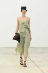 Model walking in a pale green layered slip dress with thin straps, wearing a black hat and black lace-up sandals, carrying a dark clutch on the runway