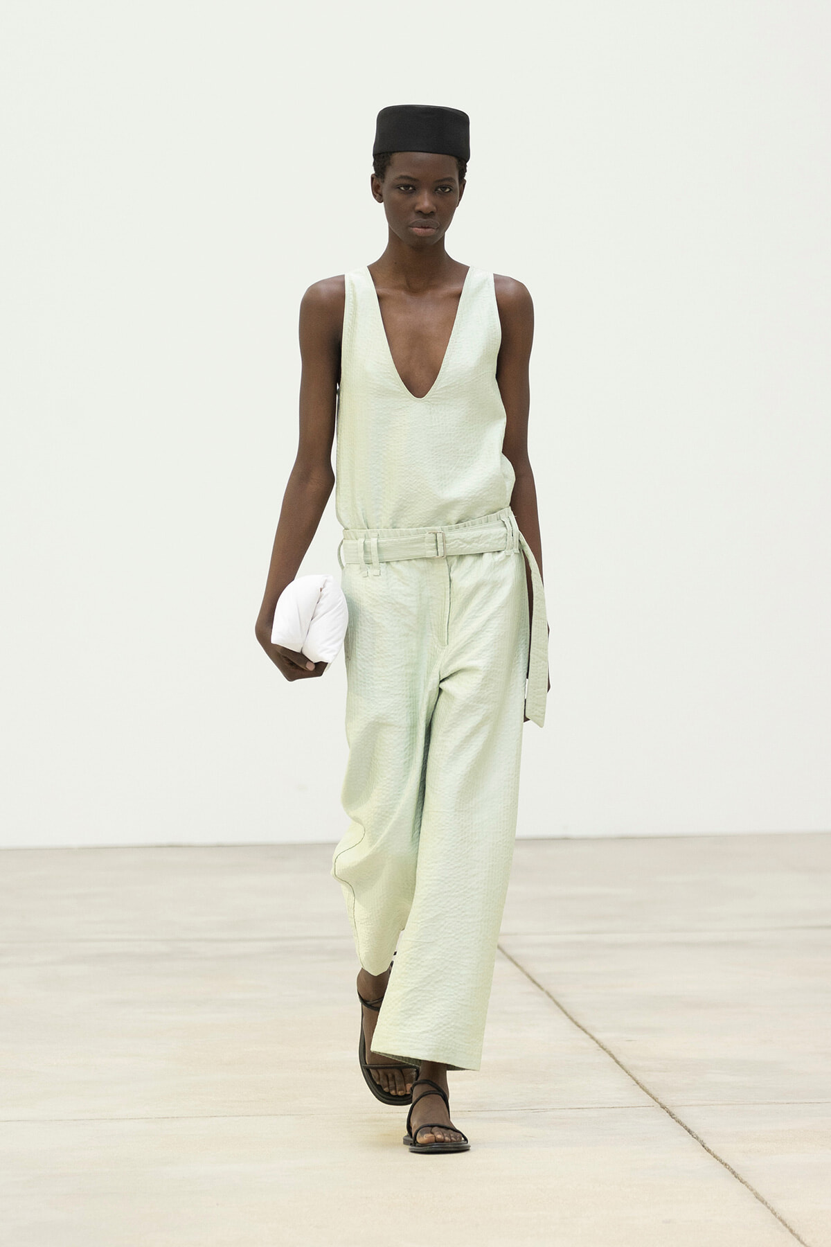 Runway model in a pale green sleeveless jumpsuit with a deep V neckline, black hat, belt, flat sandals, and a white clutch.