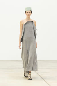 Runway model in a light gray, off-shoulder, layered dress with thin straps, paired with black strappy sandals and a mint-green cap.