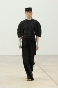 Model in a black avant-garde outfit with layered sleeves, wide-leg pants, a small black cap, large silver earrings, and a mint clutch walking on a runway.