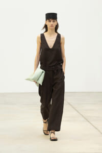 Fashion model walks in a black sleeveless V-neck top and wide black pants, belted, with a black hat and mint green clutch on a runway.