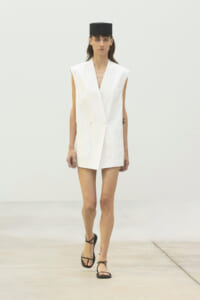 Runway model wearing a white sleeveless blazer dress with a black square hat and black strappy sandals.