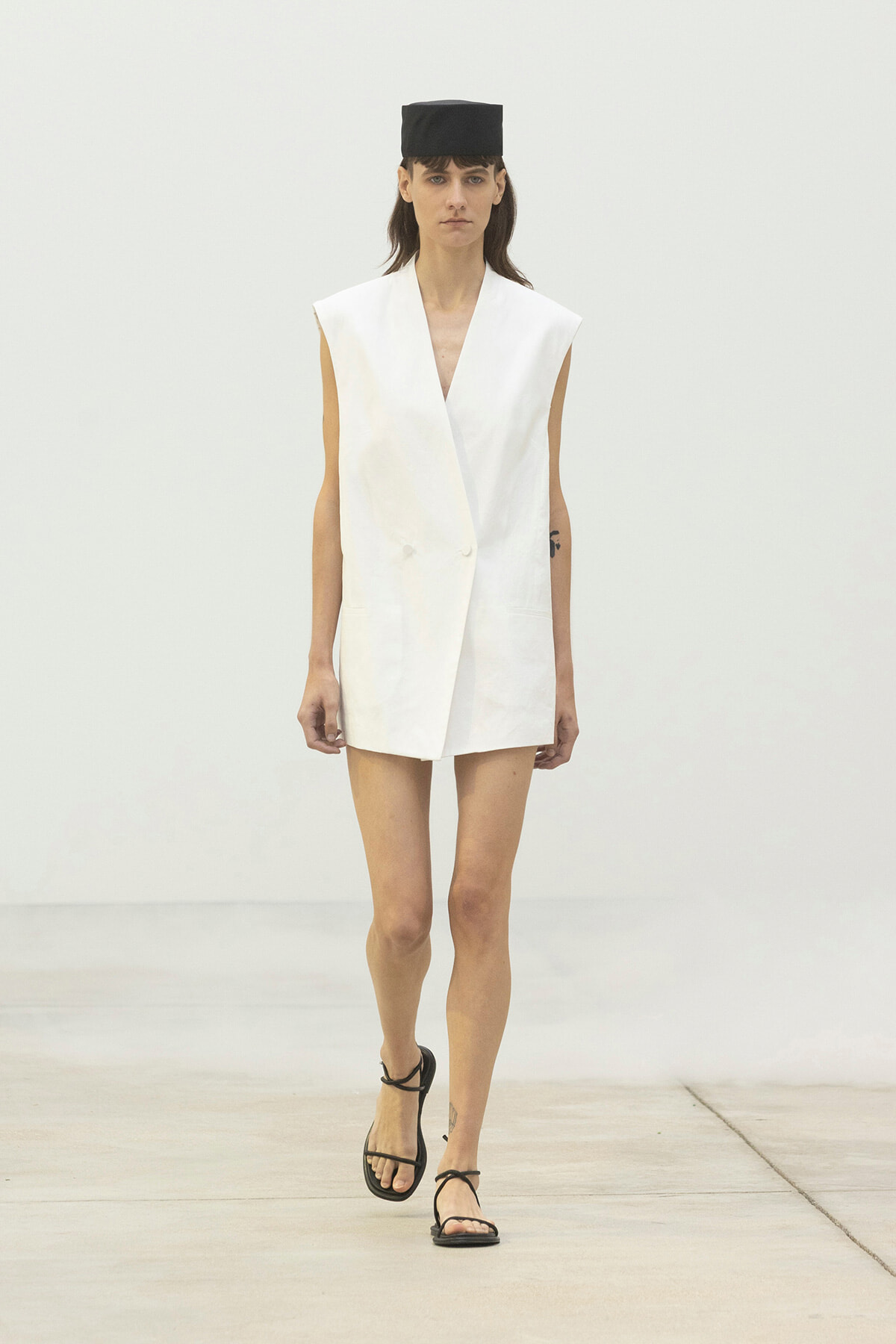 Runway model wearing a white sleeveless blazer dress with a black square hat and black strappy sandals.