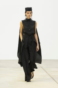 Model walking a runway in an all-black, sleeveless button-up top with a flowing cape and wide-leg belted pants, wearing a black cap and statement earrings.