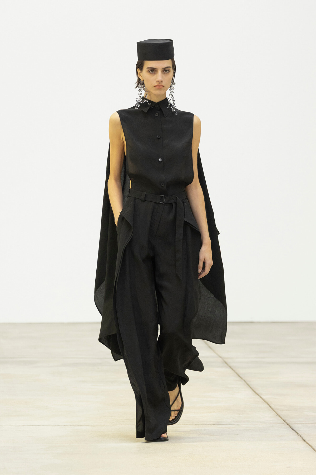 Model walking a runway in an all-black, sleeveless button-up top with a flowing cape and wide-leg belted pants, wearing a black cap and statement earrings.