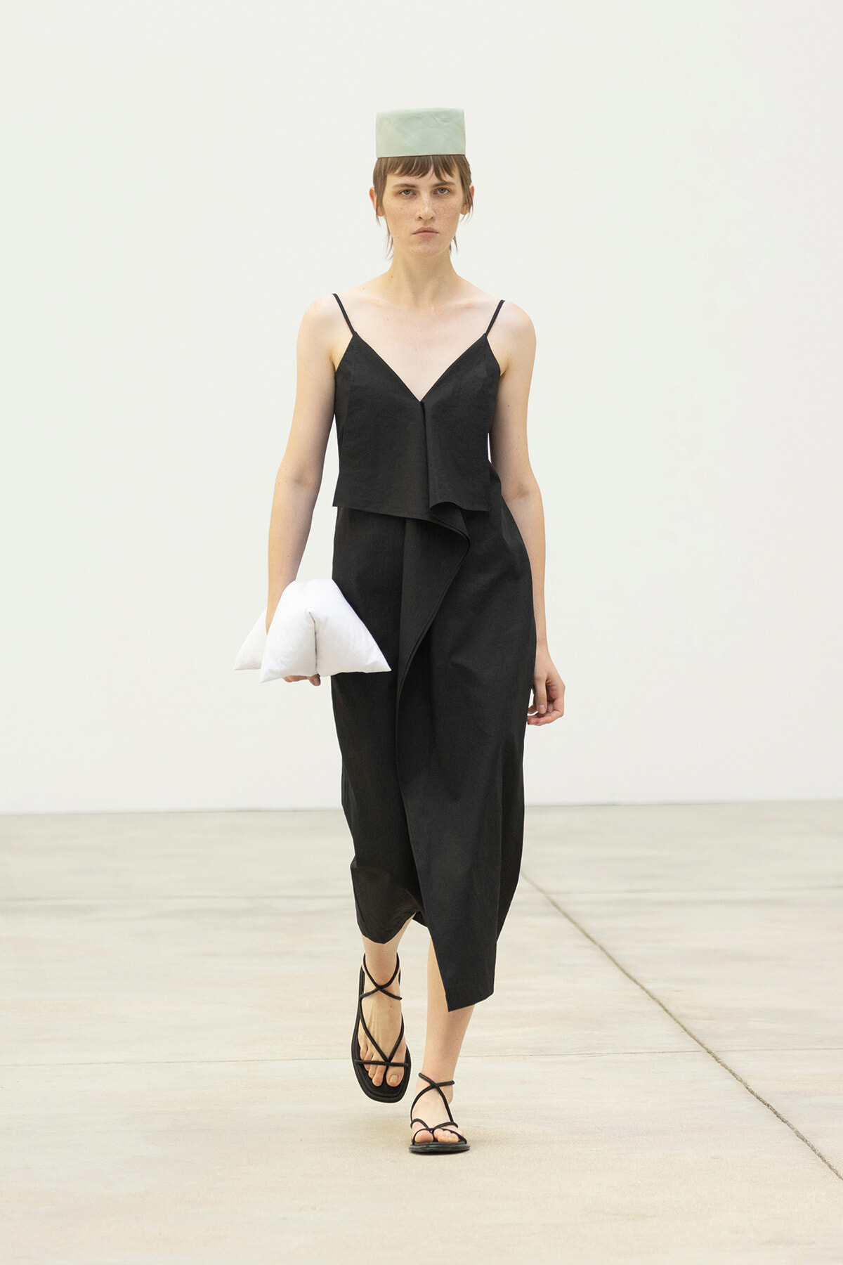 Model walking a runway in a black, layered midi dress with thin straps, a pale green square hat, and black strappy sandals, holding a white pillow-like clutch.