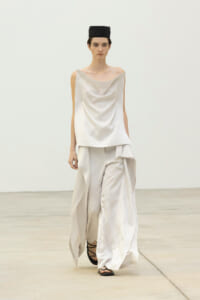 Model walks a runway in a loose, draped white top and wide-leg white pants, with a small black hat.