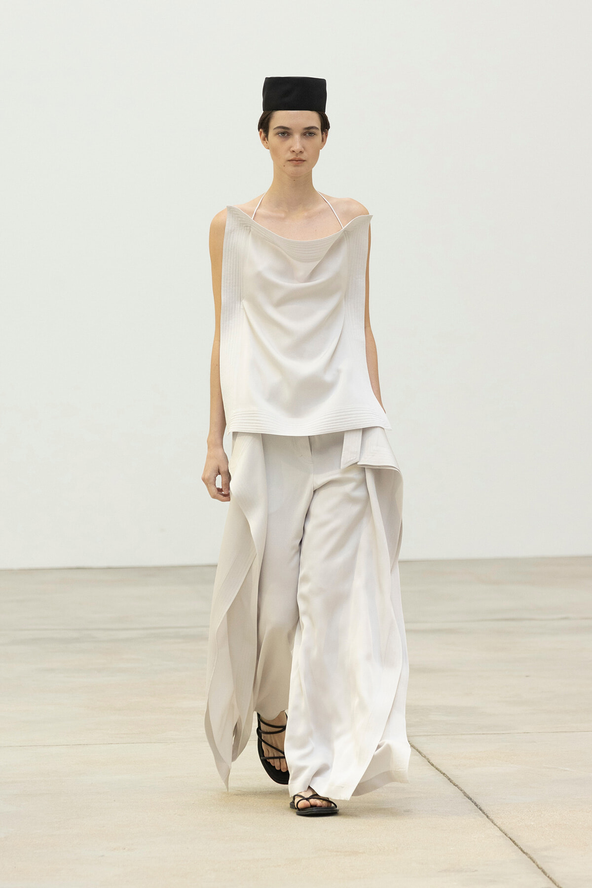 Model walks a runway in a loose, draped white top and wide-leg white pants, with a small black hat.