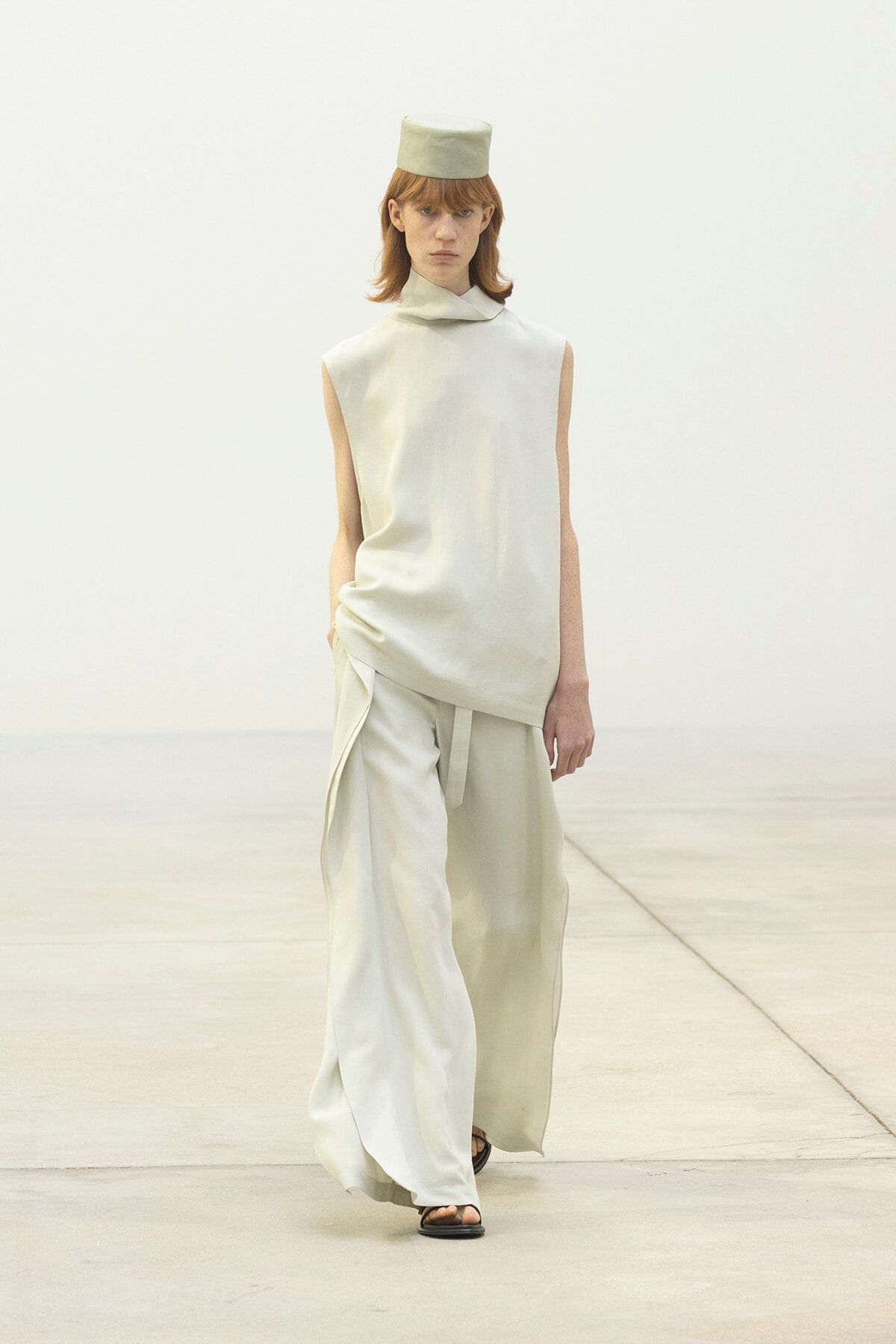 Model walking a runway in an all-cream ensemble: sleeveless draped top, wide-leg pants, and a matching hat with flat top, black sandals.