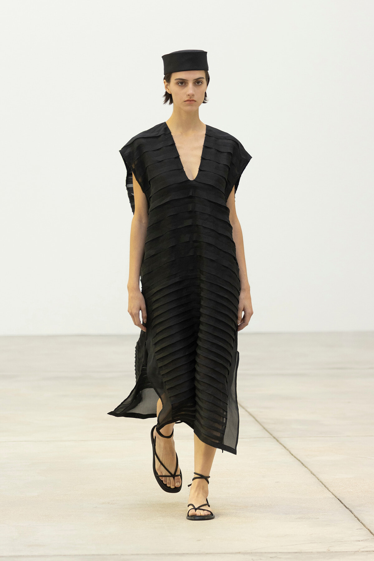 Model in a black pleated V-neck dress and cap walking on a runway, minimalist background, strappy sandals visible.