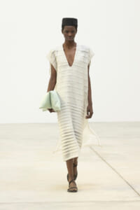 Model walking a minimalist runway in a cream, striped V-neck midi dress with a black cap and brown sandals, carrying a pale mint clutch redressed in her left hand.