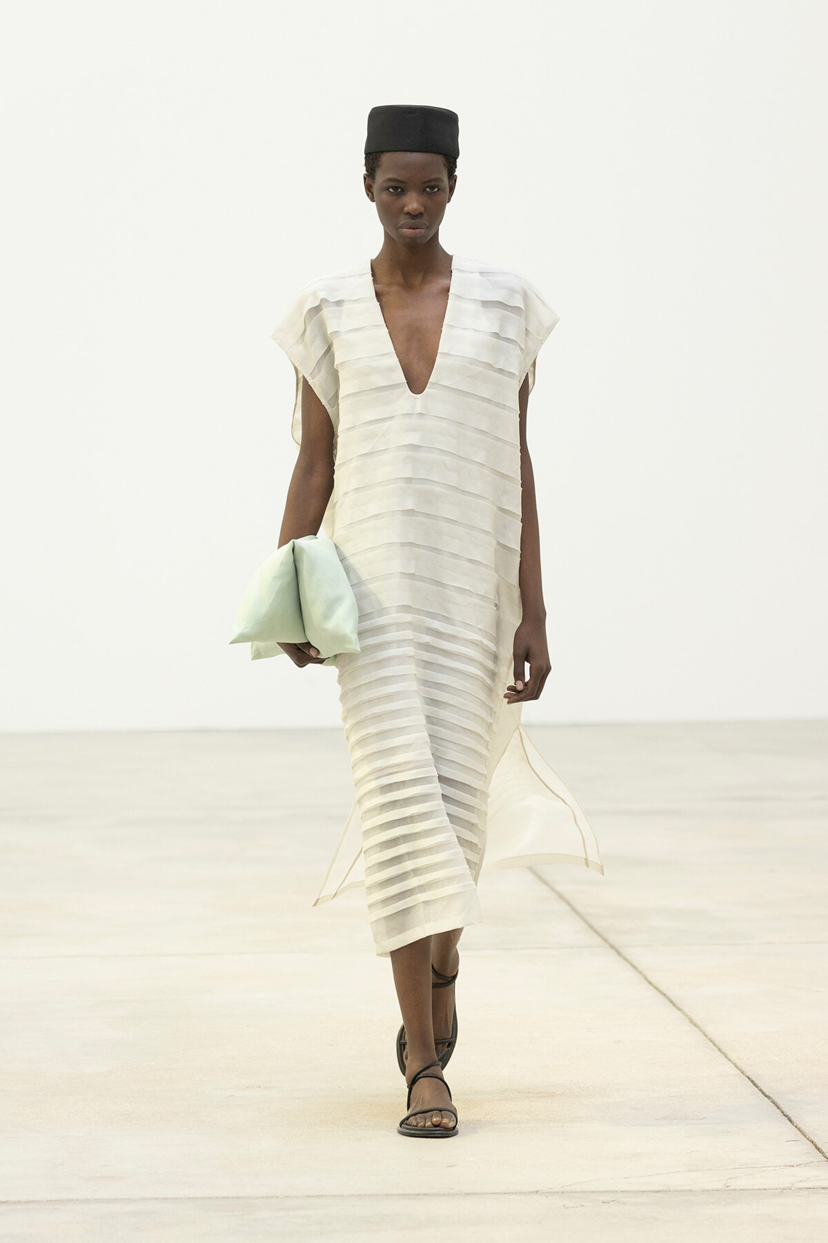 Model walking a minimalist runway in a cream, striped V-neck midi dress with a black cap and brown sandals, carrying a pale mint clutch redressed in her left hand.