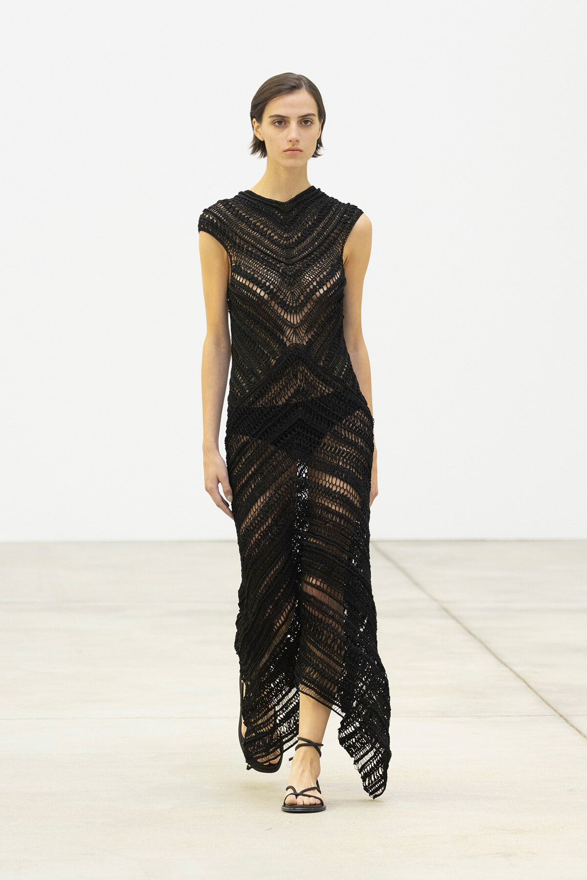 Model walking in a floor-length black crochet dress with a chevron pattern on a minimalist runway with strappy sandals.
