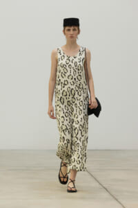 Model walking the runway in a sleeveless leopard-print jumpsuit with a black cap, black sandals, and a small black bag over her shoulder.