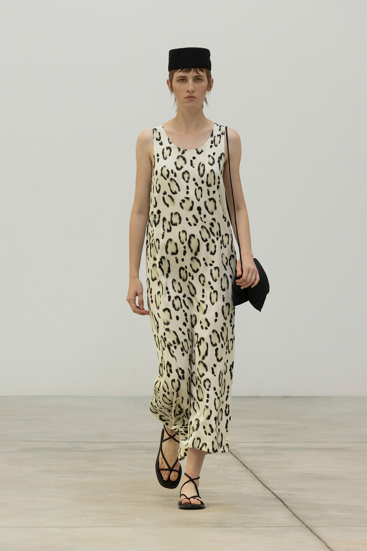 Model walking the runway in a sleeveless leopard-print jumpsuit with a black cap, black sandals, and a small black bag over her shoulder.