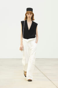 Model walks a runway wearing a black sleeveless blazer, white wide-leg cargo pants, a black hat, and strappy sandals with a small shoulder bag