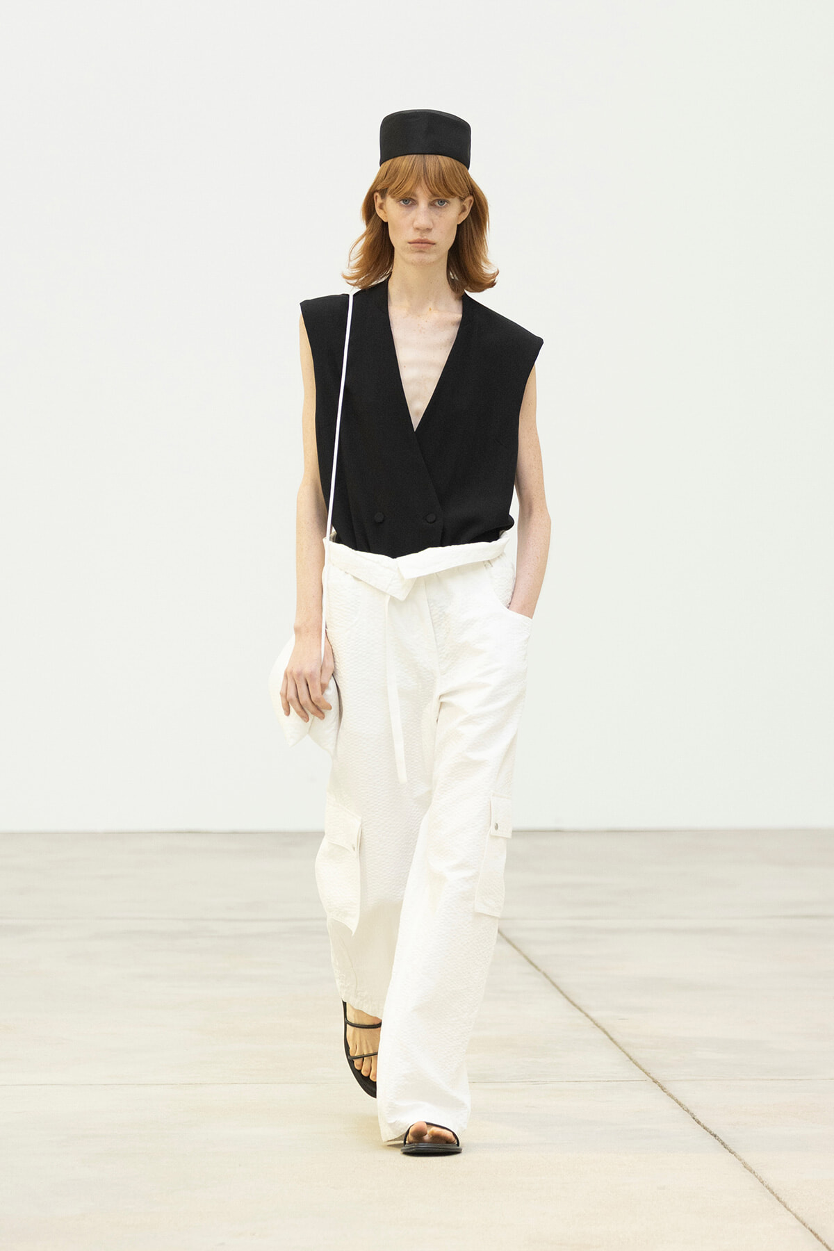 Model walks a runway wearing a black sleeveless blazer, white wide-leg cargo pants, a black hat, and strappy sandals with a small shoulder bag