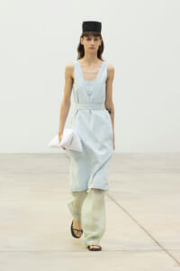 Runway model in a light blue sleeveless dress belted at the waist, over pale green wide‑leg pants, with a black cap, black sandals, and a white clutch held in her right hand.